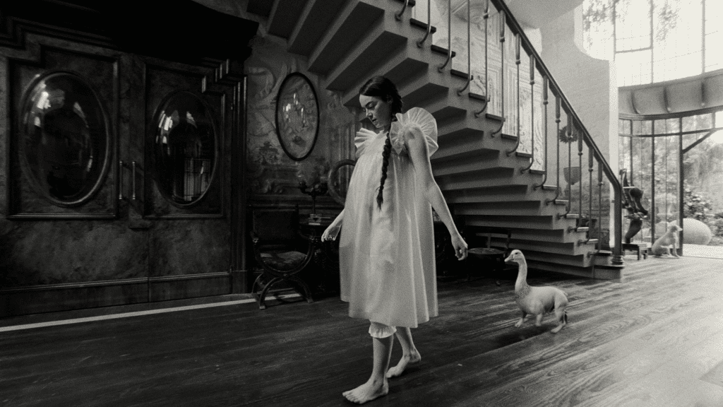 Bella Baxter (Emma Stone) au sein du manoir londonien de son maître, Godwin Baxter , toute vêtue de blanc. Derrière elle, suivant ses pas, un animal hybride, entre l'oie et le chien. Image en noir et blanc, issue des trente premières minutes du film, Pauvres Créatures. © 2023 Searchlight Pictures All Rights Reserved.