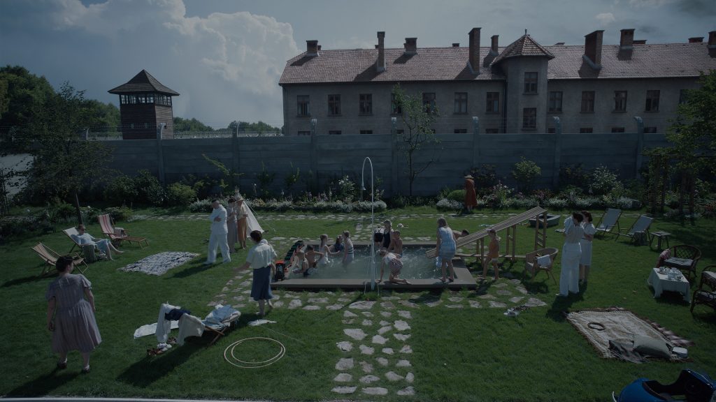 Plan sur le jardin de la famille Höss, avec la piscine, les enfants qui jouent, les invités qui profitent du temps radieux, avec le mur du camp en fond et des bâtiment d’Auschwitz visibles, au delà du mur - © Bac Films / DR